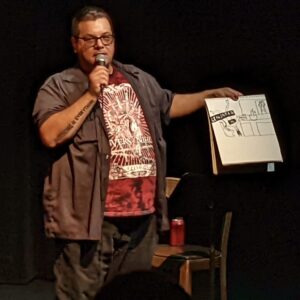 Jose Macall showing a drawing of a 911 call while talking into a microphone at The Narrators.